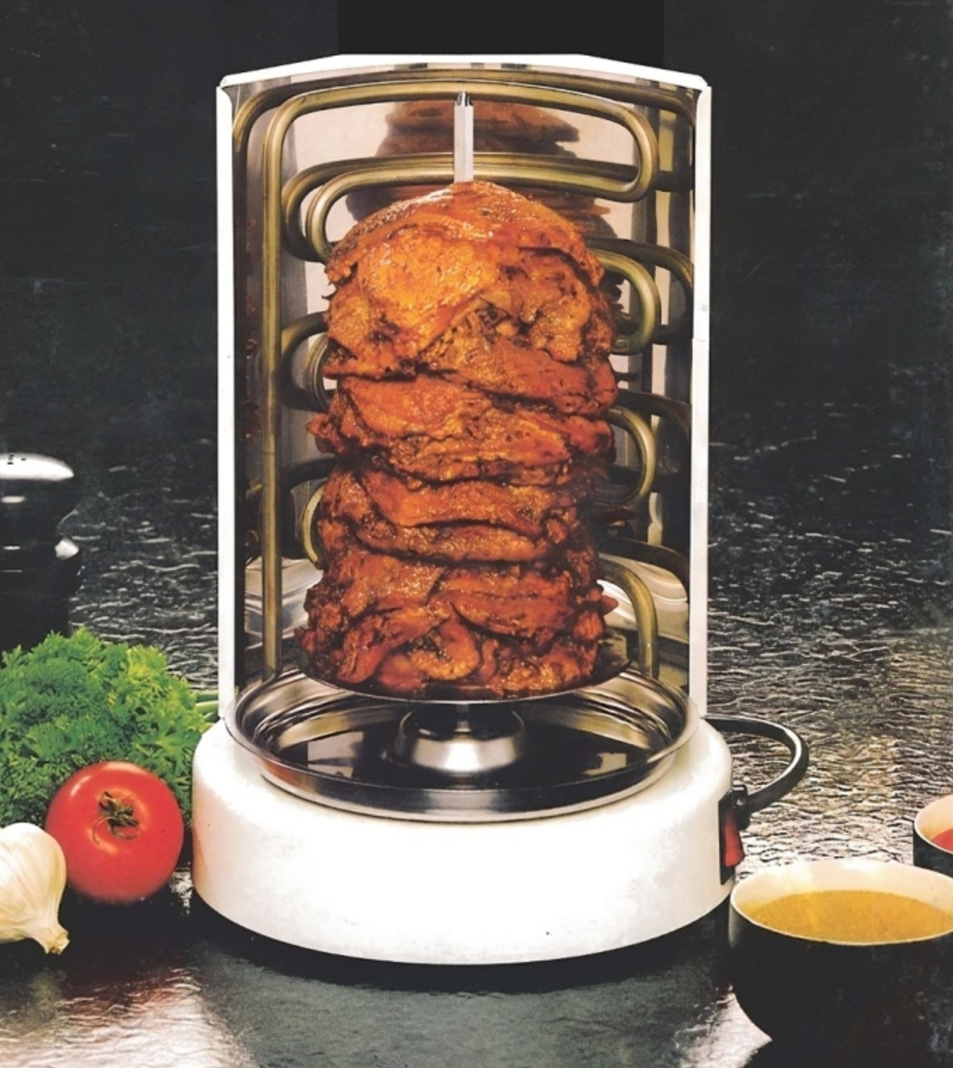 Stack of fried chicken on a vertical rotisserie grill with vegetables and dipping sauce on a dark surface.
